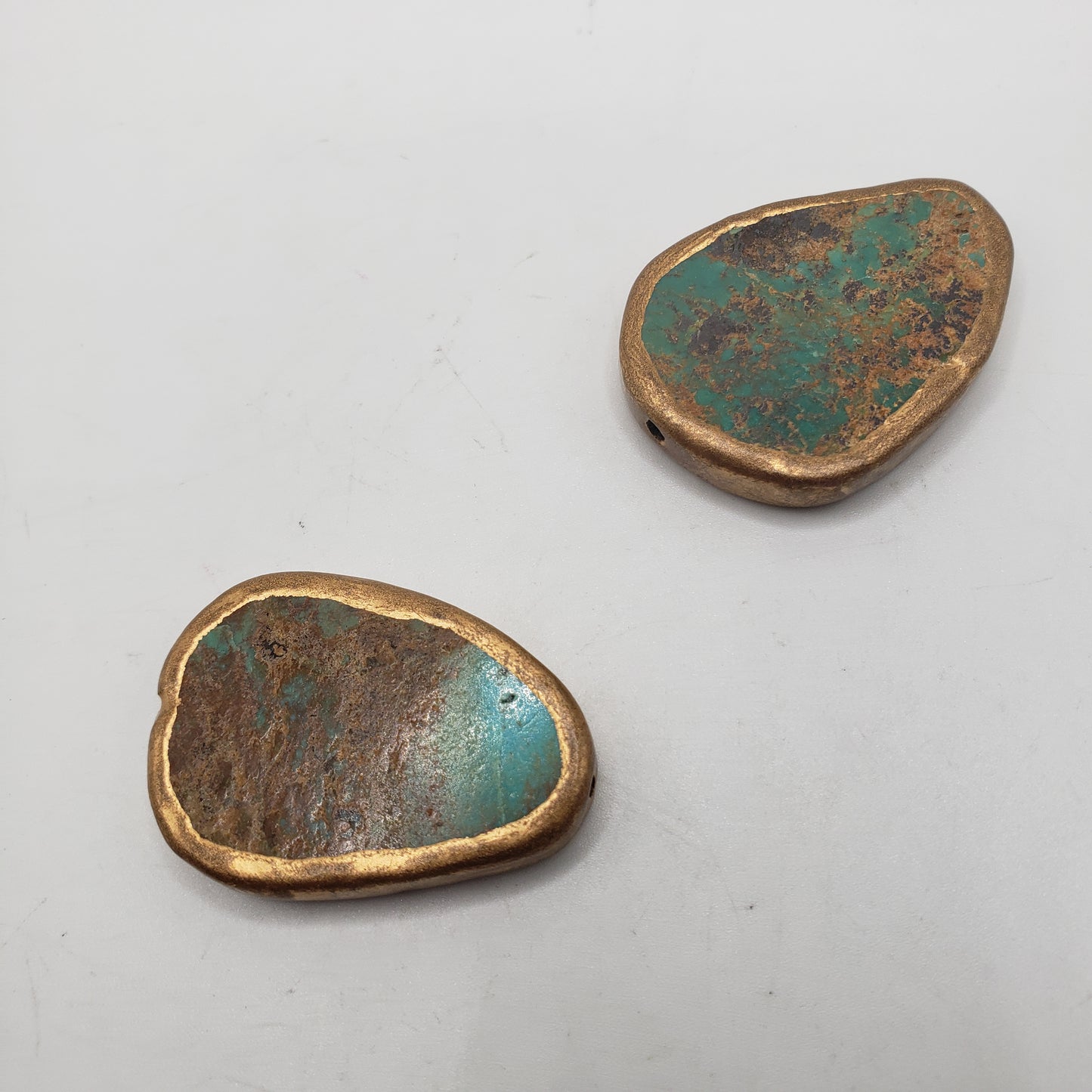 Two Copper Plated Turquoise Single stones with irregular shapes. The stones feature a blend of greenish-blue and brown tones, along with a rough texture and metallic edges. They are placed on a smooth, light-colored surface.