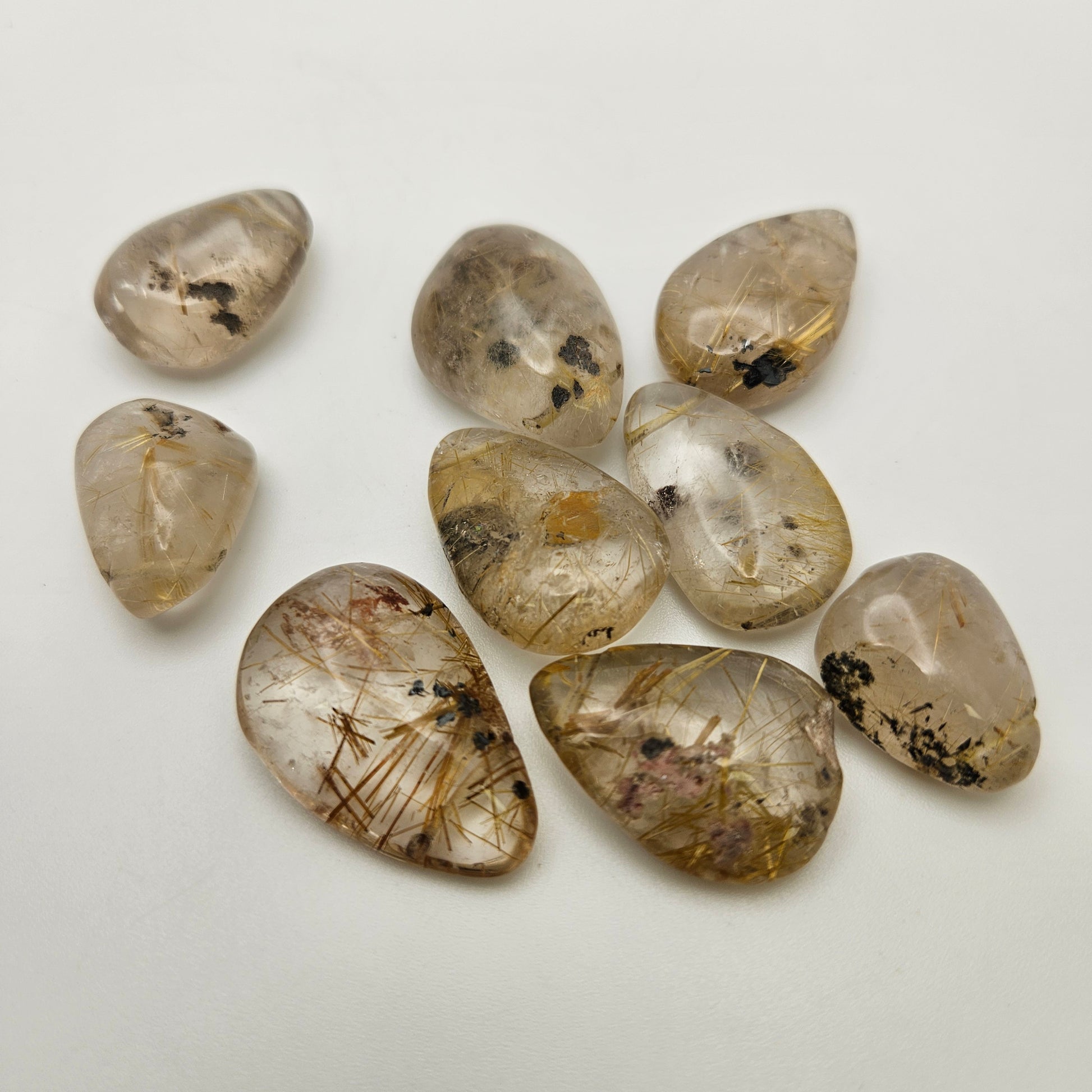 Nine Assorted Gold Rutilated Quartz Top Drill SINGLE stones, featuring polished surfaces and golden needle-like inclusions, are arranged on a white background. Each stone is tear-shaped and varies in size, with unique patterns and occasional black flecks.