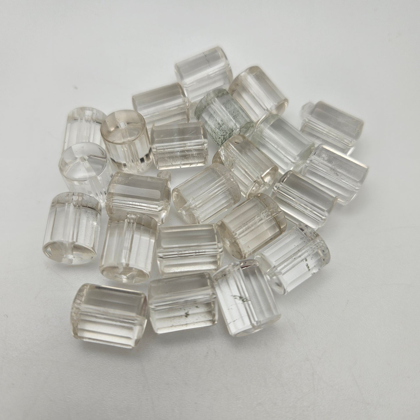 A collection of 20mm Clear Quartz Tube SINGLE beads is scattered on a plain white surface. The tubes are transparent and exhibit subtle variations, displaying slight imperfections and internal reflections.