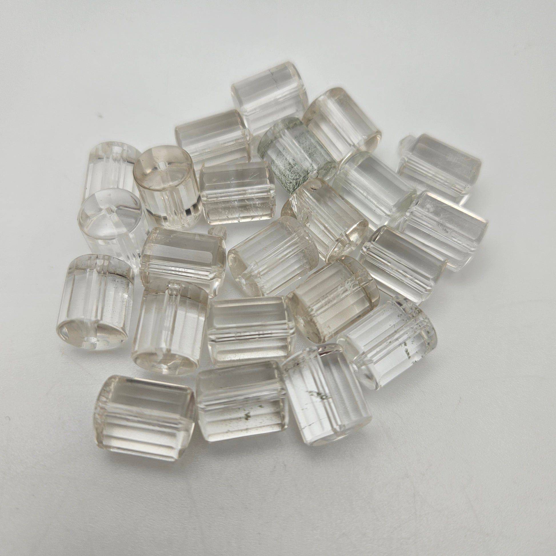 A collection of 20mm Clear Quartz Tube SINGLE beads is scattered on a plain white surface. The tubes are transparent and exhibit subtle variations, displaying slight imperfections and internal reflections.