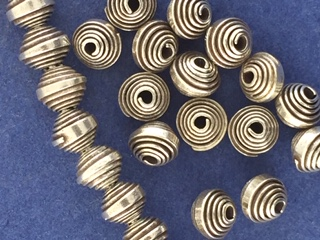 Close-up of sterling silver small round spacer beads with spiral decoration, artistically arranged on a blue surface. The 6mm wide beads vary slightly in size and display intricate spiral patterns. A partial strand begins on the left, curving gracefully, while others are scattered across the right side.