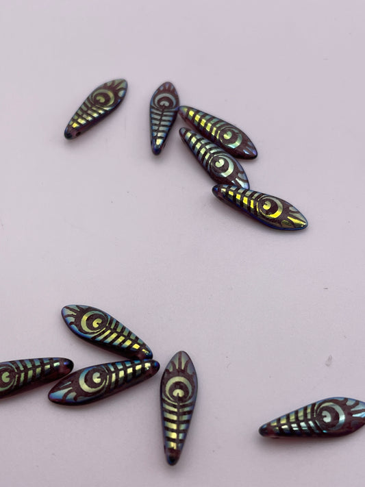 A variety of 15mm Peacock Leaf Pattern Spear Top Drilled Czech Single Beads are displayed on a light surface. These iridescent beads feature intricate eye-like patterns and a glossy finish that reflects different colors. Some beads are grouped together, while others are scattered around.