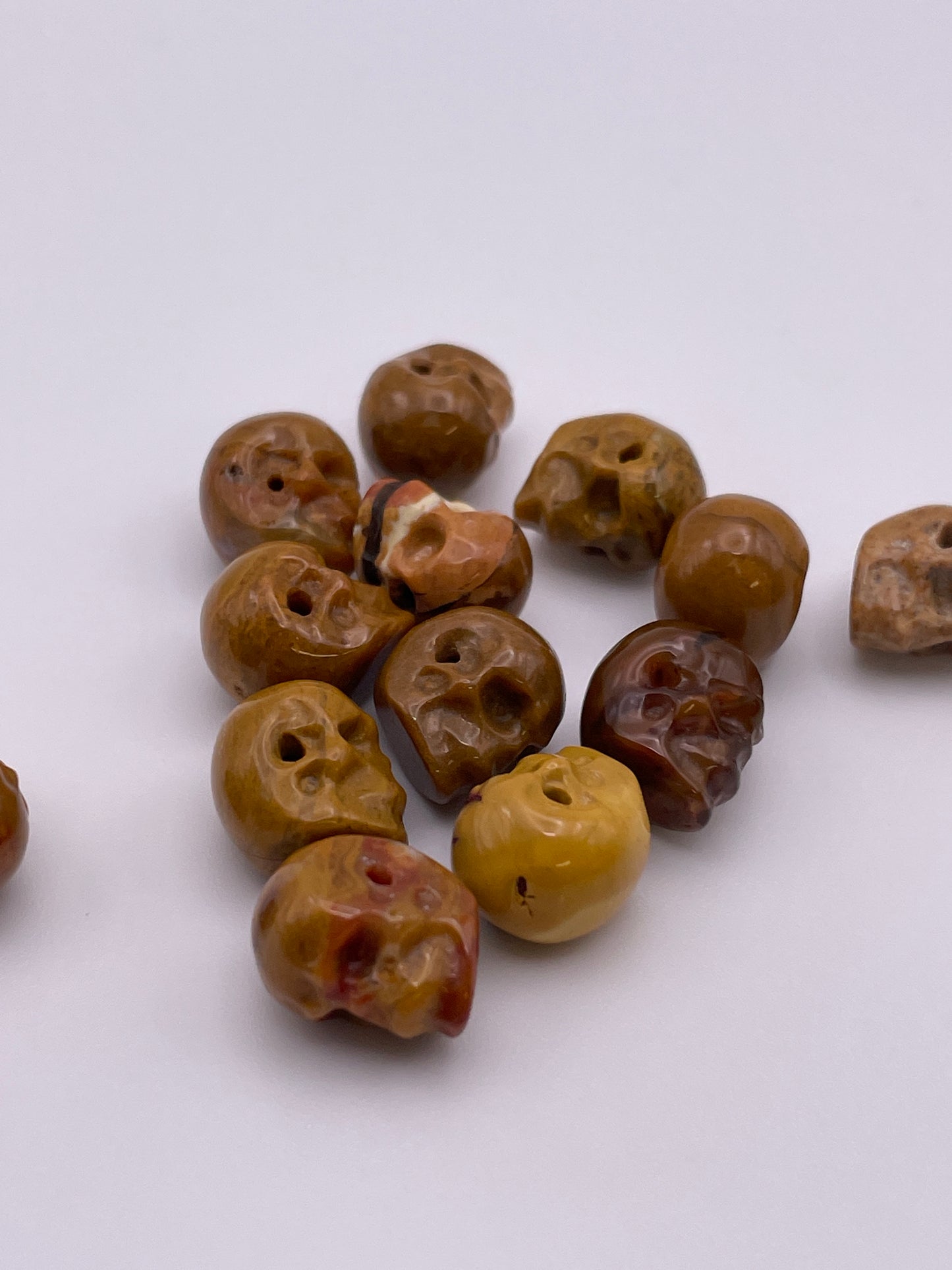 On a plain white surface lies an array of 8mm Mookaite Skull Single Beads. Each polished, skull-shaped bead displays unique variations in color and texture, contributing to their rustic appearance.