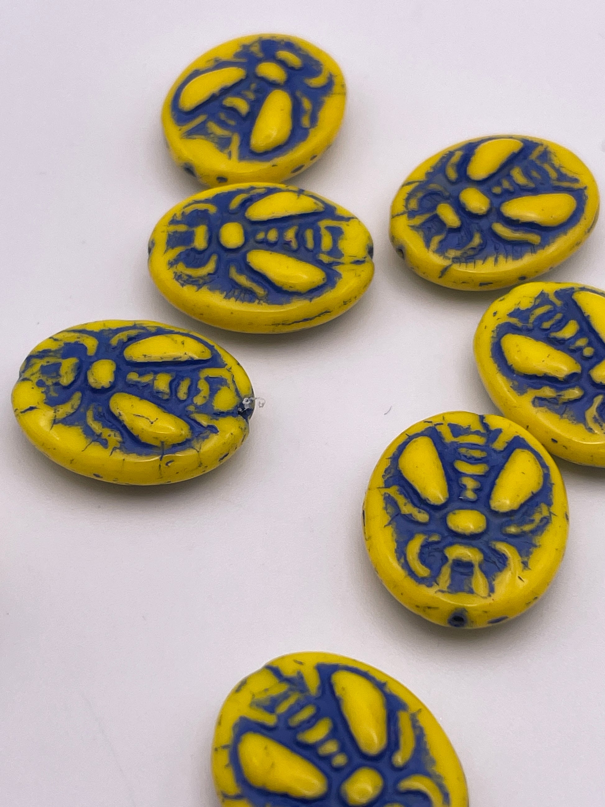 Seven 20mm Yellow & Blue Bee Oval Czech Beads are scattered on a white surface.