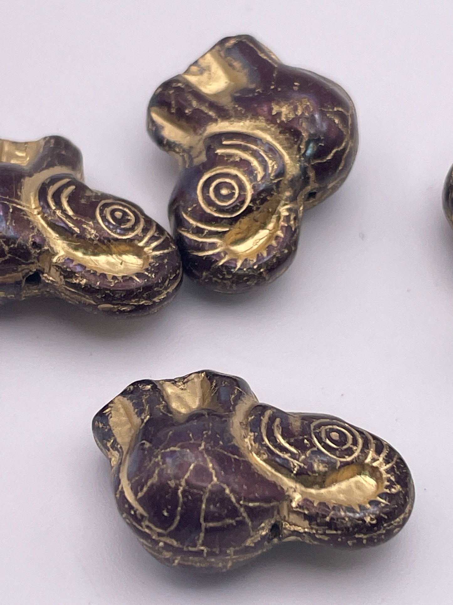 Close-up of several 20mm Dark Metallic Elephant Czech Single Beads with intricate detailing, including engraved spirals on the ears. The beads have a worn effect, highlighting their metallic finish against a plain white background.